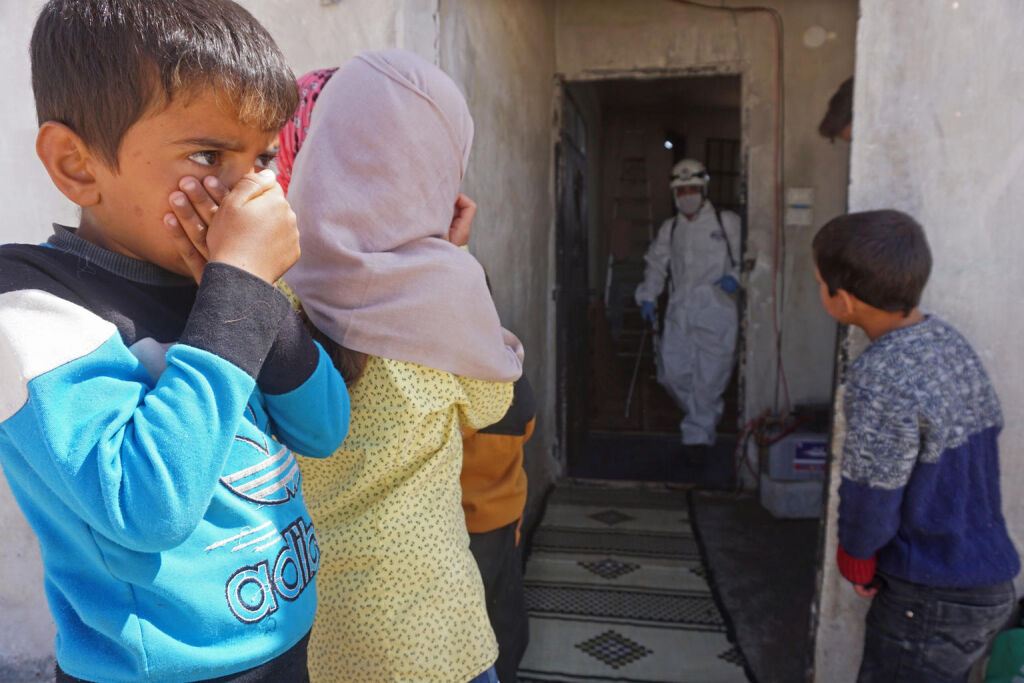 Lapset seuraavat, kuinka entistä koulurakennusta desinfioidaan maaliskuun 26. päivä Binnishin kaupungissa Syyriassa. Rakennus toimii nyt maansisäisten pakolaisten hätämajoituksena. © UNICEF/UNI316131/Haj Kadour/AFP