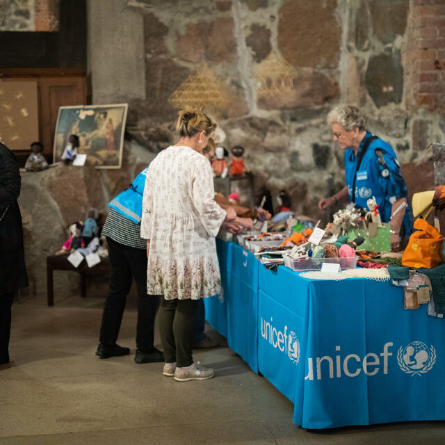 Myyntipöytä, jossa UNICEFIn logolla varustettu pöytäliina ja paljon myyntituotteita. Ihmisiä katselee tuotteita.