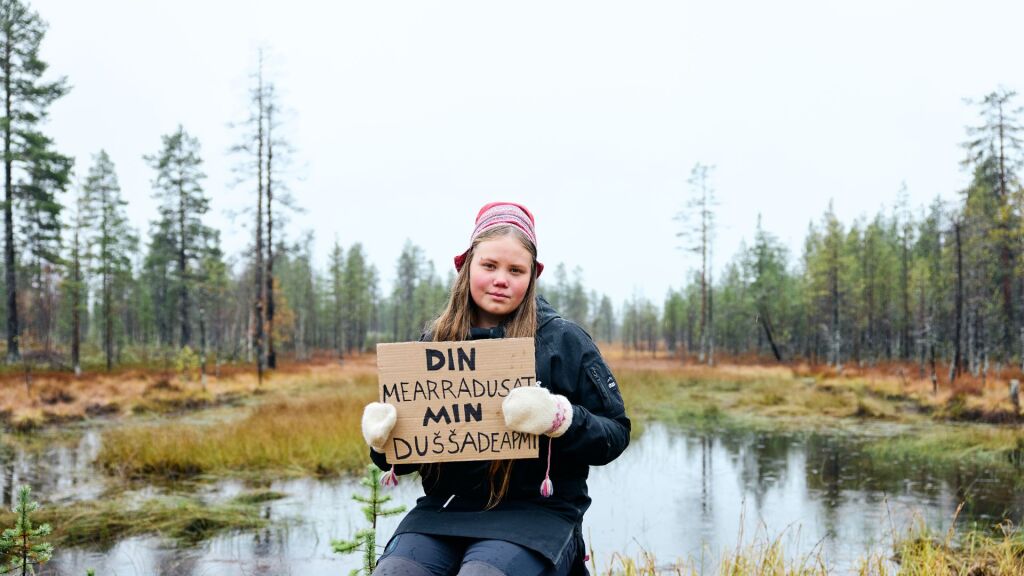 Nuori istuu jakkaralla kyltti kädessään sateisessa syksyisessä maisemassa pienen lammen edessä, Kyltissä on saamenkielistä kirjoitusta. 