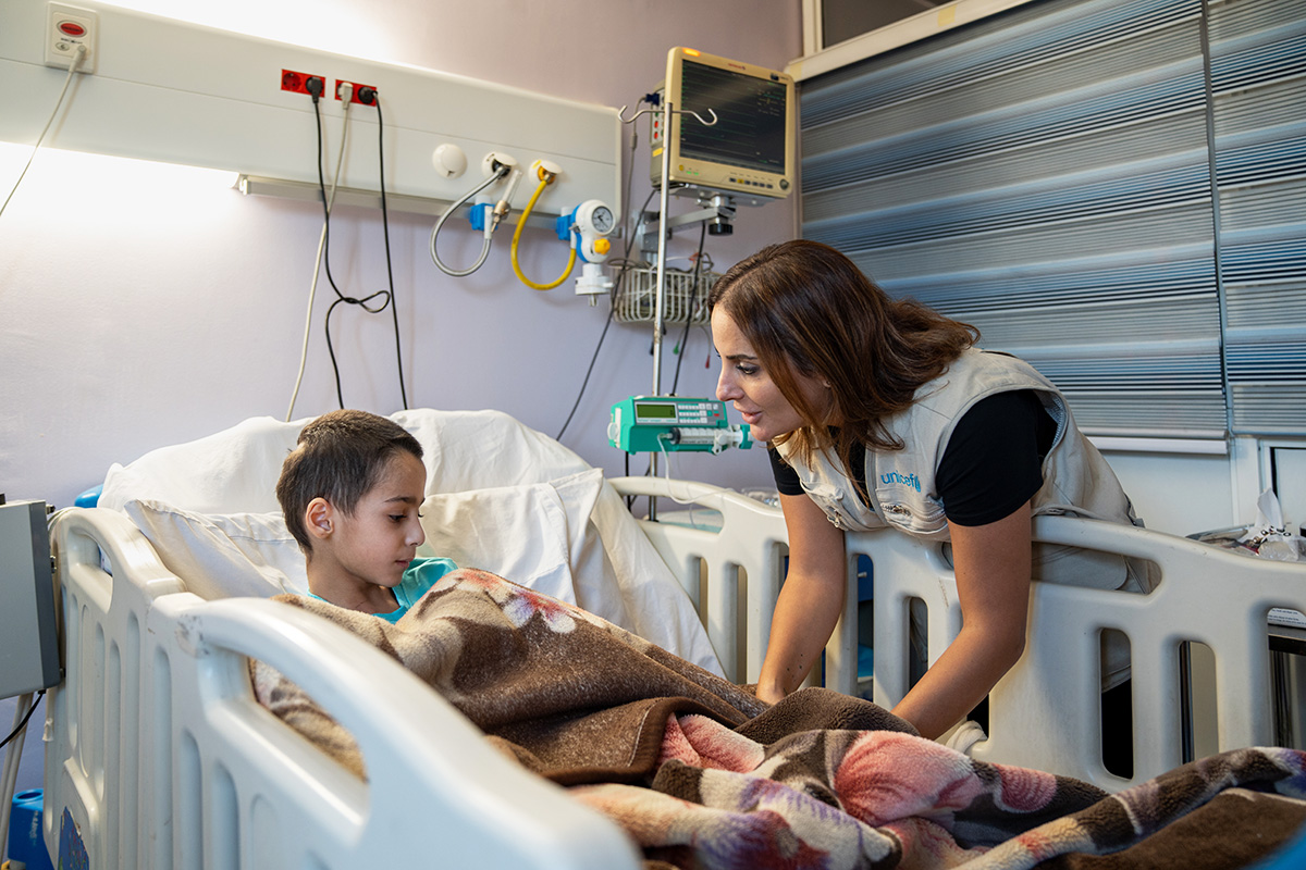 Lapsi, jolla takaraivossa haava, makaa sairaalasängyssä. UNICEFin työntekijä juttelee lapselle. 