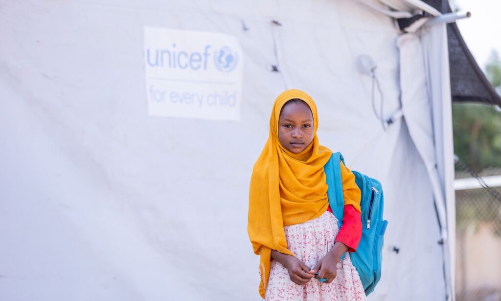 Lapsi UNICEF-teltan edessä koulureppu selässään.