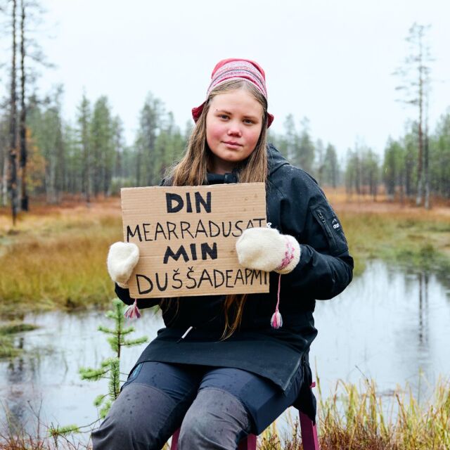 Nuori istuu jakkaralla kyltti kädessään sateisessa syksyisessä maisemassa pienen lammen edessä, Kyltissä on saamenkielistä kirjoitusta. 