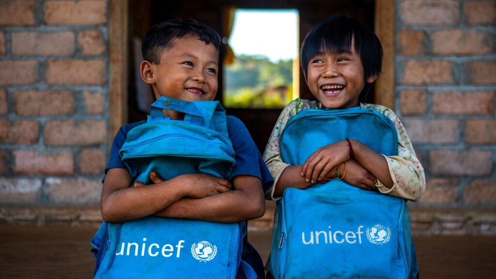 Kaksi nauravaa alakouluikäistä lasta istuu vierekkäin ja puristaa sylissään likaantuneita sinisiä koulureppuja, joissa on UNICEFin logo. 