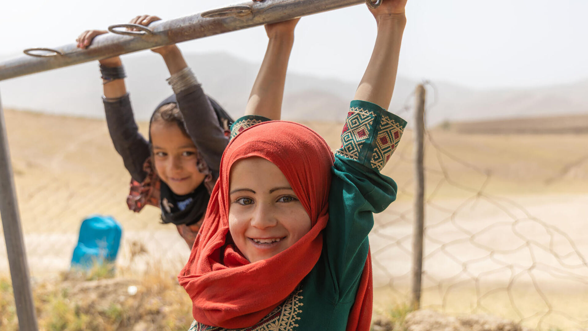 Kaksi alakouluikäistä huiveihin pukeutunutta lasta keinuu kiipeilytelineessä.