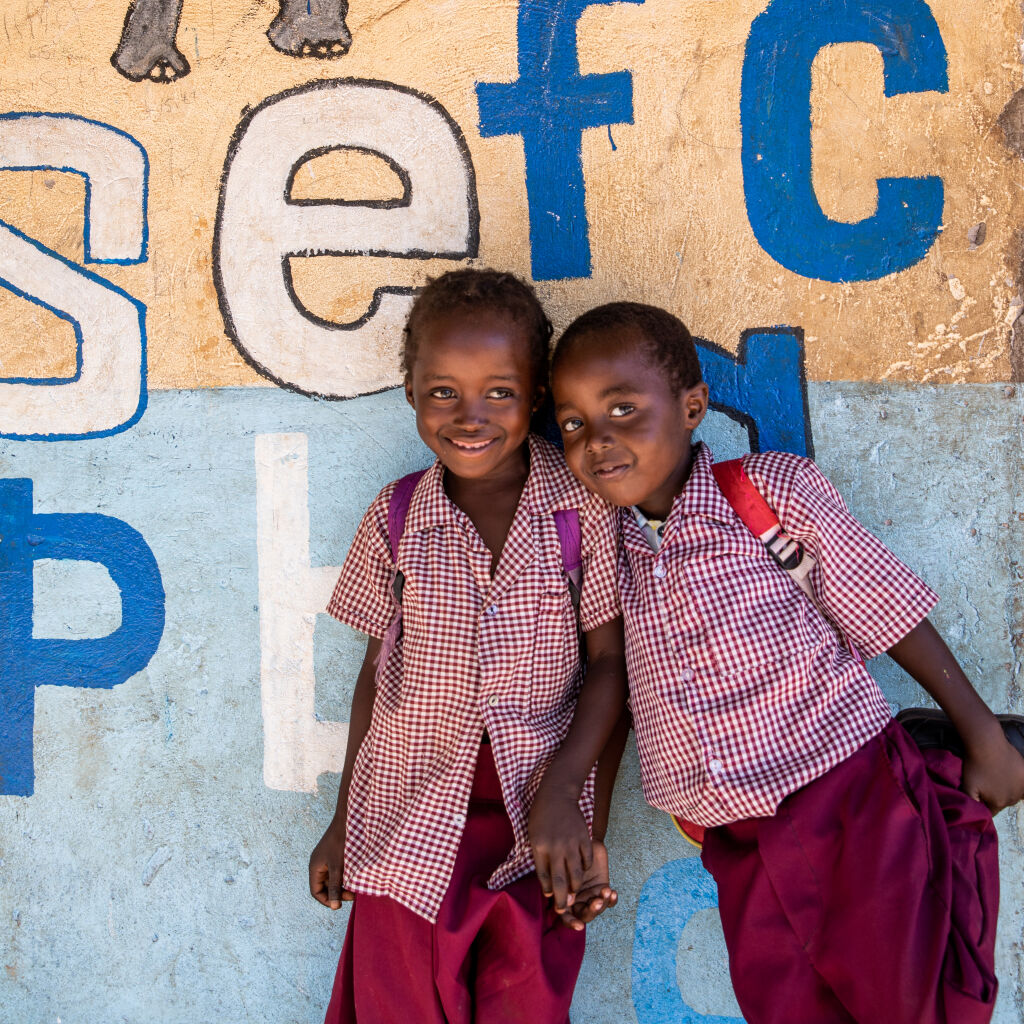 Two young schoolchildren leaning against a wall that has some of the alphabet drawn on it