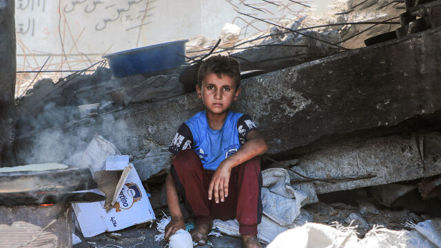 Lapsi istuu tuhoutuneen rakennuksen edessä ja katsoo vakavana kameraan. 
