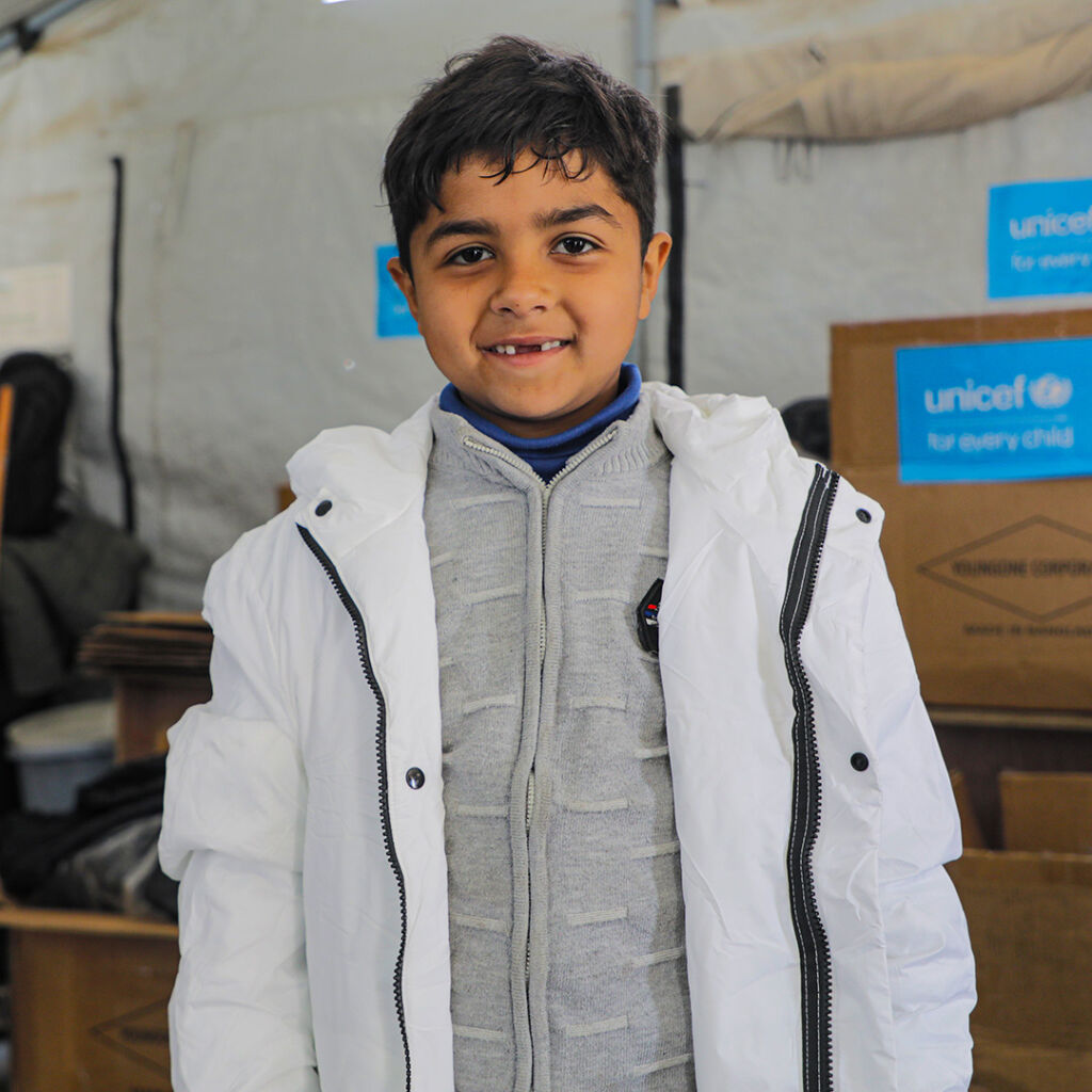 Lapsi seisoo teltassa päällään upouusi talvitakki ja hymyilee leveästi. Taustalla pahvilaatikkoja, joissa UNICEFn logo.