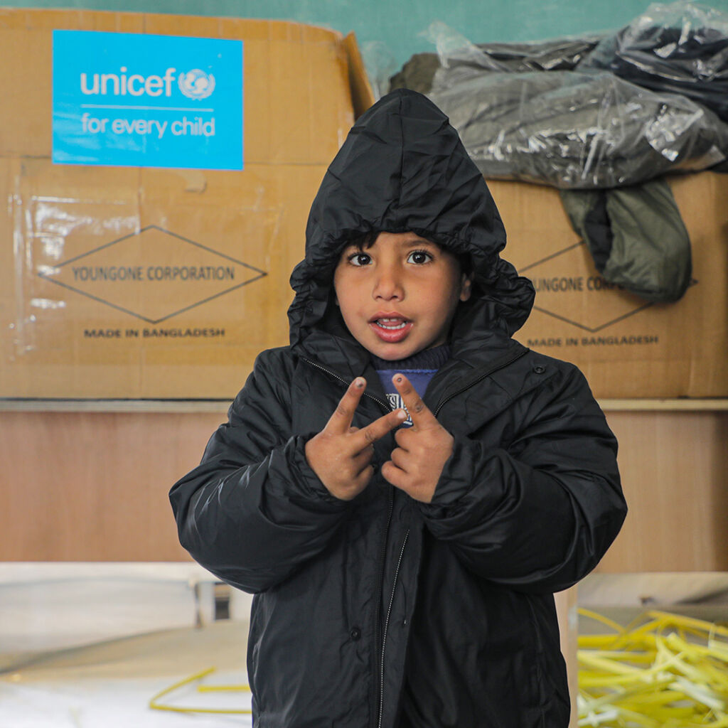 Lapsi päällään talvitakki seisoo teltassa ja hymyilee kameralle. Taustalla pahvilaatikoita, joissa UNICEFin logo.
