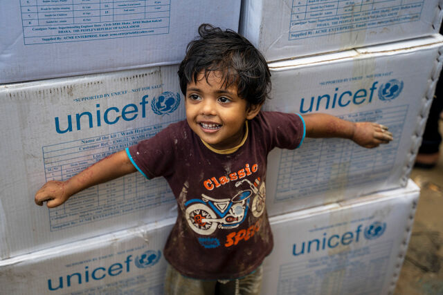 Lapsi hymyilee ja seisoo valkoisten laatikoiden edessä, joissa on UNICEFin logo.