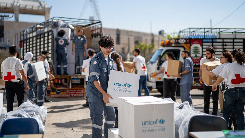 Kuvassa rekkoja, laatikoita ja avustustyöntekijöitä. Mies kantaa laatikkoa, jossa lukee UNICEF.