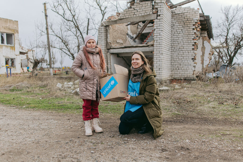 Lapsi ja UNICEFin työntekijä tuhoutuneen rakennuksen edessä.