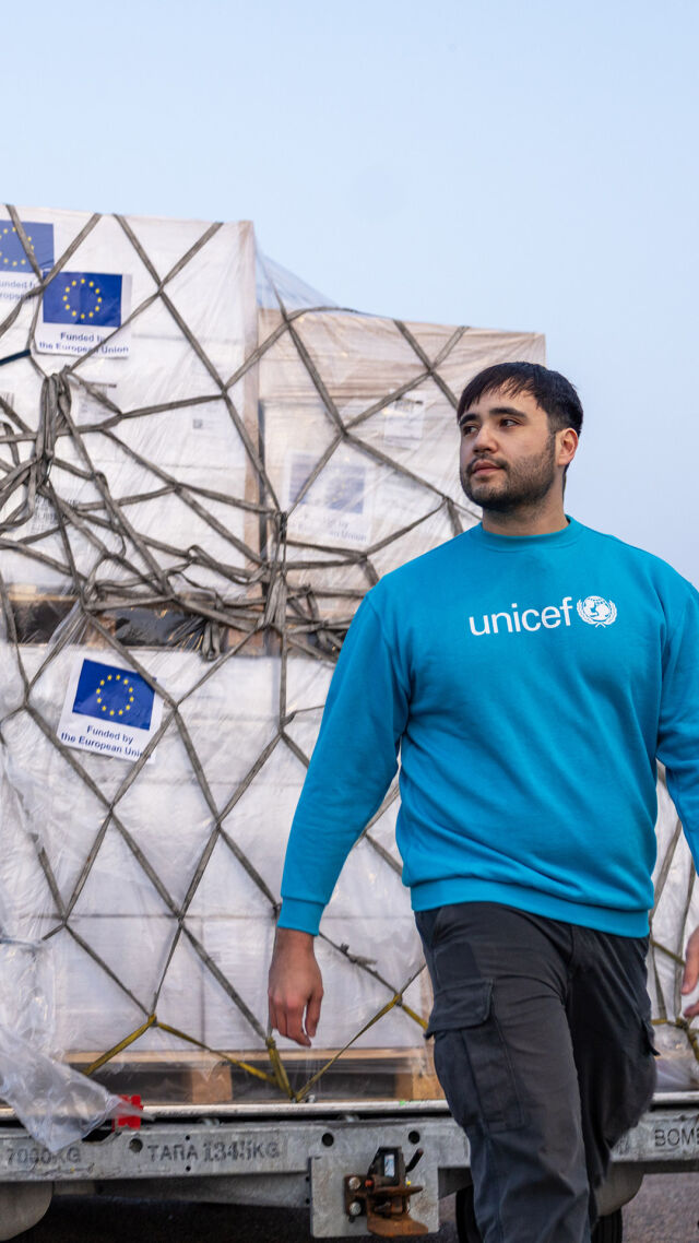Lentokenttä, jolla isoja laatikkokasoja, joissa UNICEFin ja EU:n tarrat. UNICEFin työntekijä edustalla. 