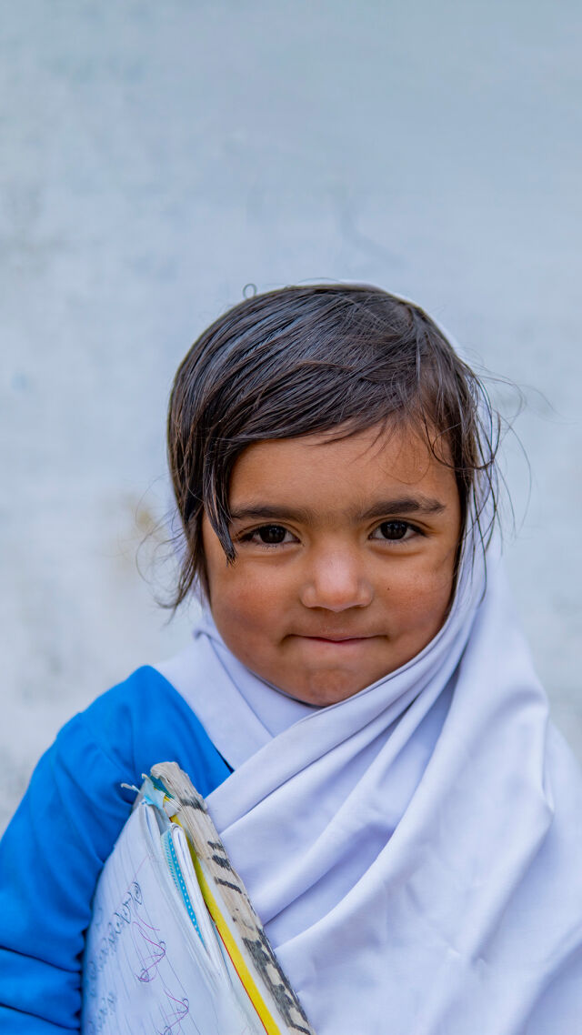 Huivipäinen tyttö kainalossaan vihko hymyilee kameralle. 