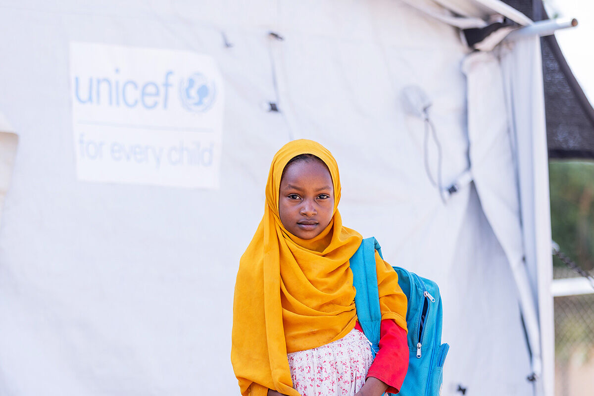 Lapsi seisoo reppu selässä teltan edessä, jonka kyljessä on UNICEFIn logo.