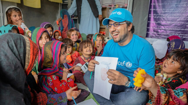 Mies kantaa laatikkoa, jossa on UNICEFin logo. Taustalla iso pino samanlaisia laatikoita. 