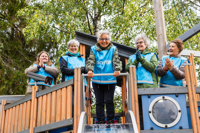 Viisi aikuista naista siniset UNICEF-liivit päällä lasten leikkitelineessä nauraa. 