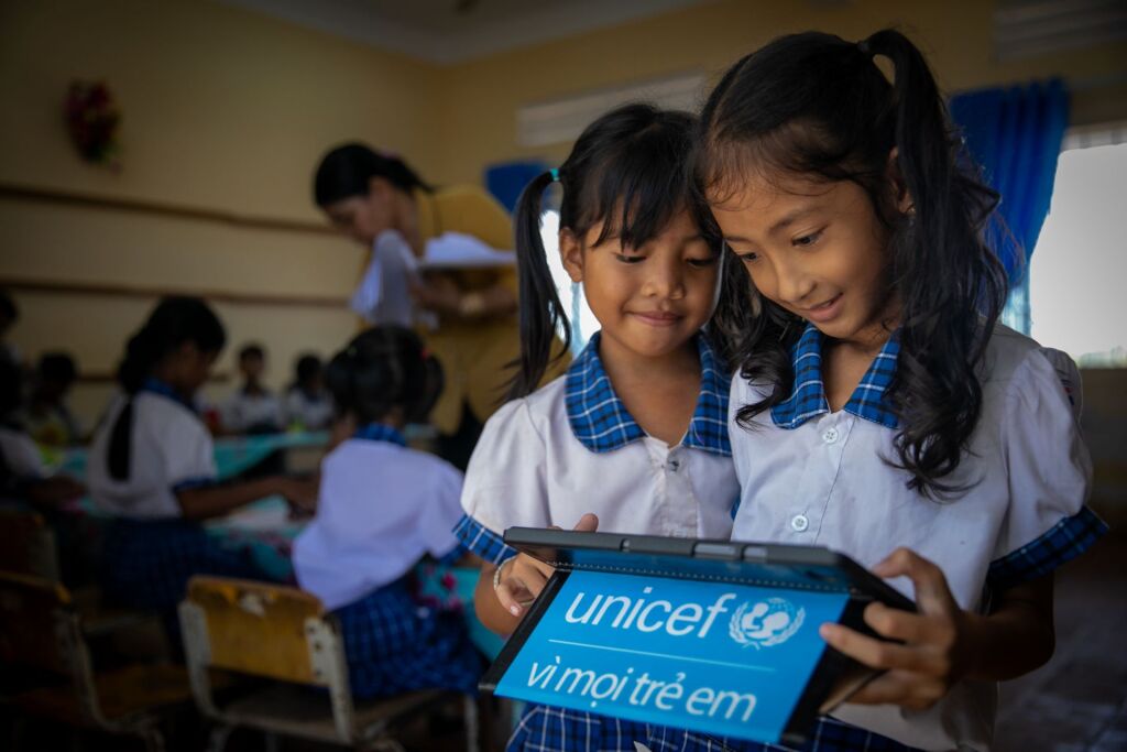 Kaksi lasta vietnamilaisessa luokkahuoneessa tutustuu UNICEFin digitaaliseen oppimateriaaliin tabletilta, opettaja taustalla.