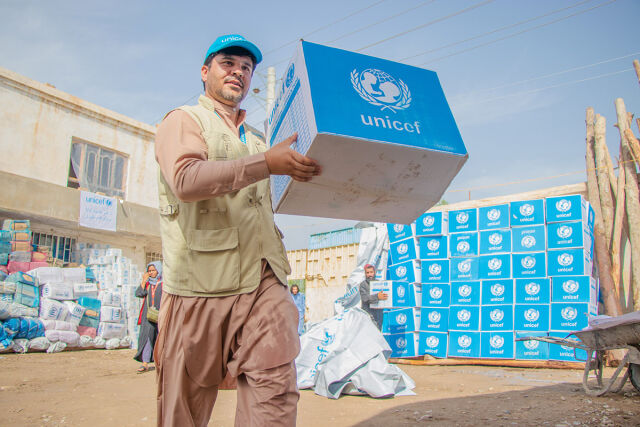 Mies kantaa laatikkoa, jossa on UNICEFin logo. Taustalla iso pino samanlaisia laatikoita.